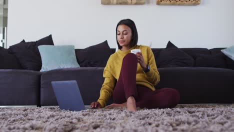 Mixed-race-woman-sitting-on-floor-using-laptop-drinking-cup-of-coffee