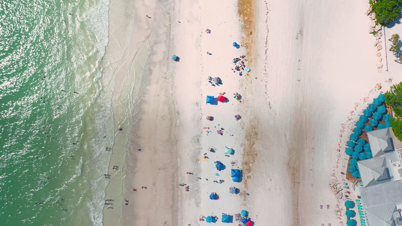 Colorful umbrellas dot the sandy shoreline as turquoise waves meet the coast, with nearby resort facilities visible in a bright aerial perspective