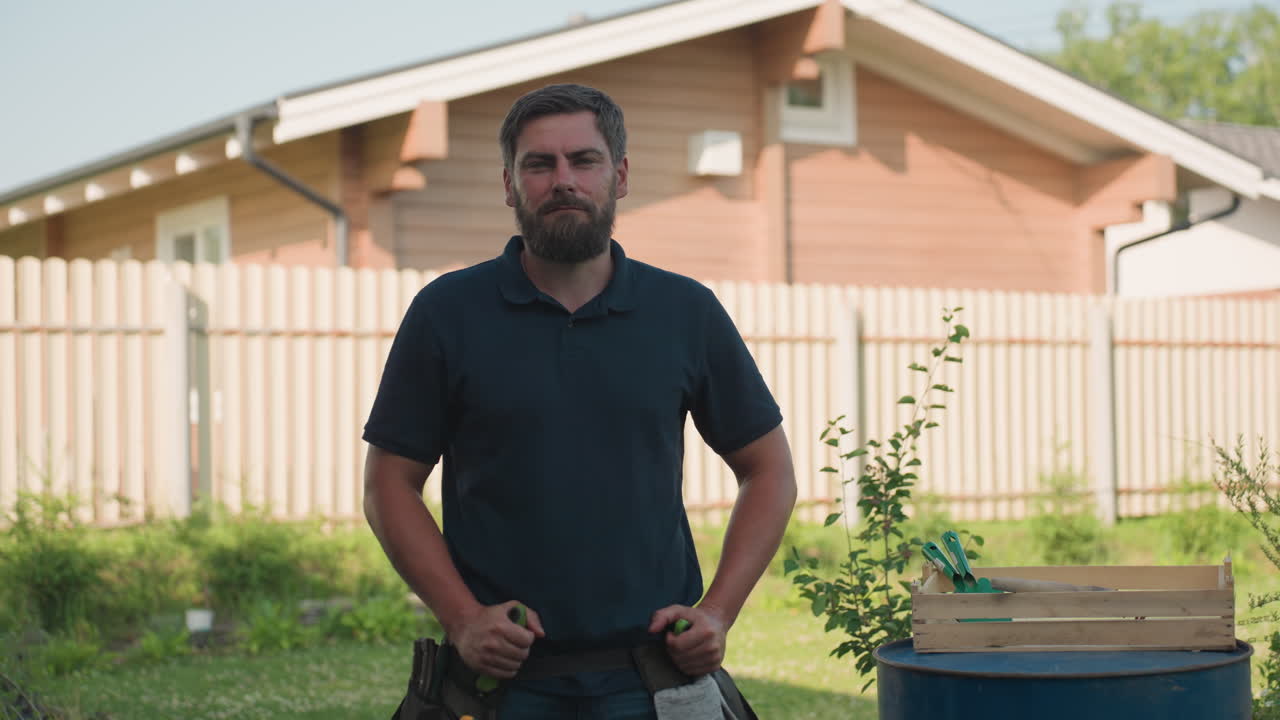 Bearded gardener in dark polo stands hands on waist, then removes hand to grip trowel from belt pocket beside blue drum with crate of tools in sunny backyard near wooden house