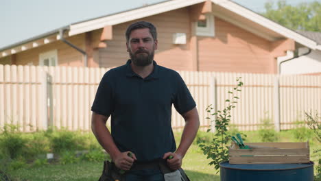 bearded gardener in dark polo stands hands on waist, then removes hand to grip trowel from belt pocket beside blue drum with crate of tools in sunny backyard near wooden house