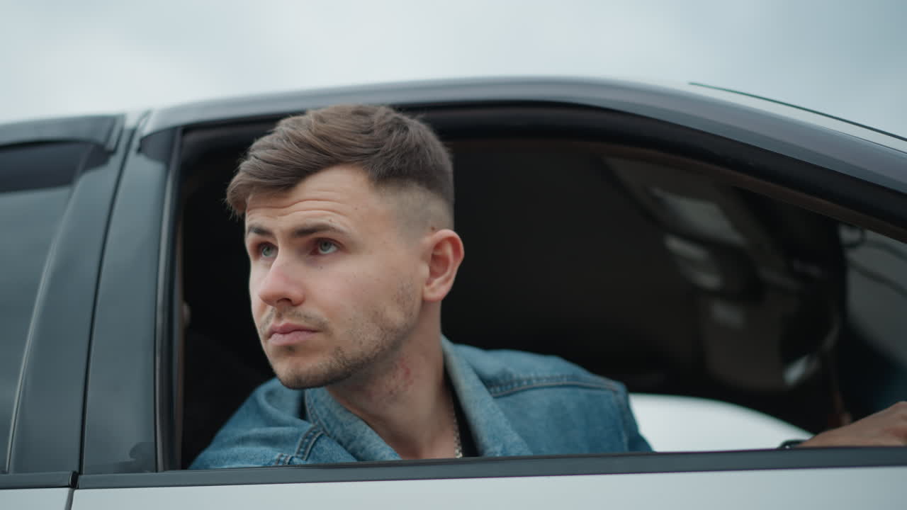 Homem branco dirigindo, debruçado na janela de um sedã branco, observando a beira da estrada e o horizonte, jaqueta jeans, retrato de perfil, checando o fluxo de tráfego e o meio-fio, contexto de estrada suburbana, expressão alerta e calma