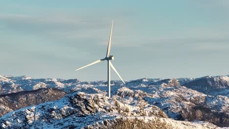 drone shot of wind turbine spinning above snow capped hills on sunny winter day