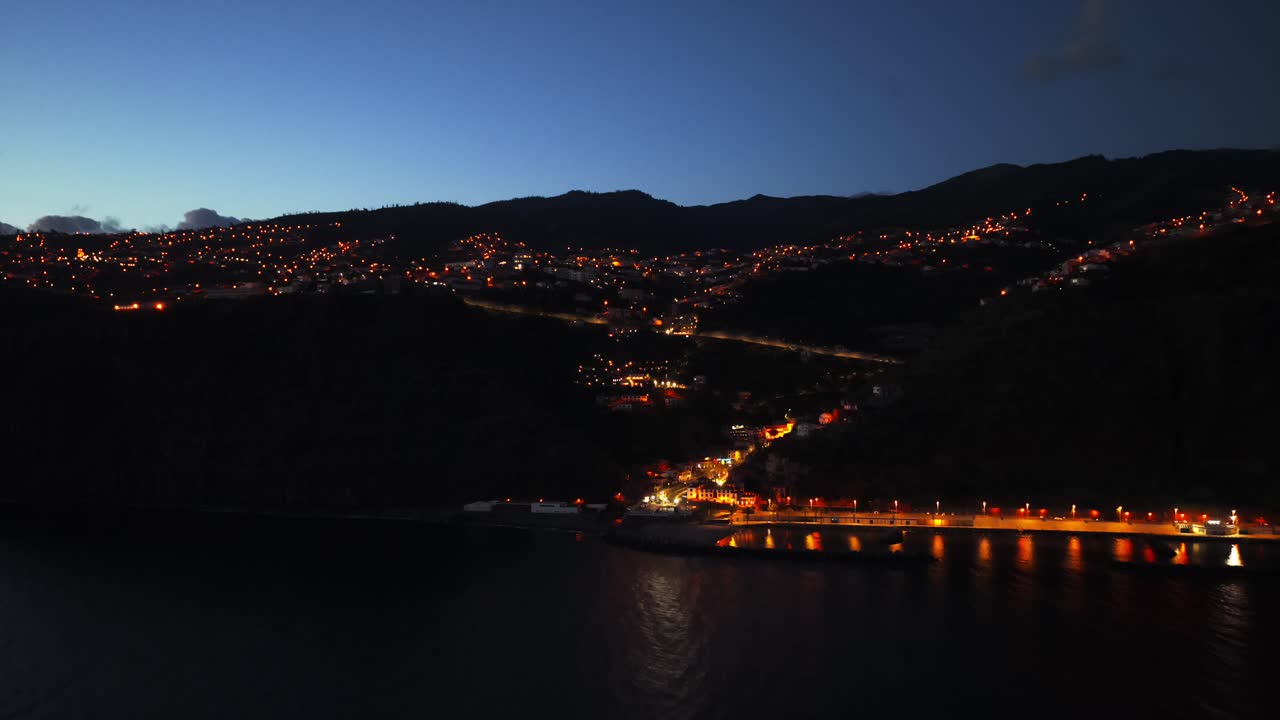 Illuminated Calheta town and harbor at twilight, city lights dot hillside by calm sea, Madeira, Portugal. Aerial forward,copy space
