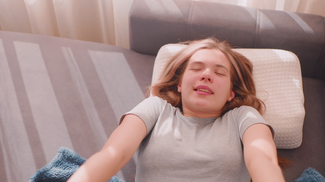 Lazy girl finally wakes up stretching herself on couch with blanket and pillow, eyes closed enjoying warmth as sunlight streams through curtains