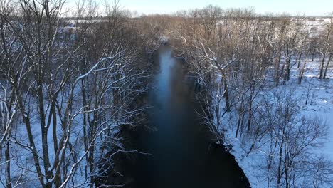 Aufsteigende-Drohnenaufnahme-Eines-Flusses-Mit-Blattlosen-Bäumen-Im-Winterschnee