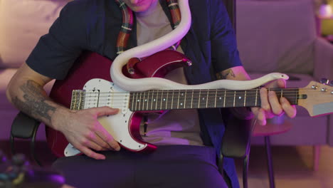 Unrecognizable-Man-Sitting-On-A-Chair-Holding-Snakes-Around-His-Neck-And-Playing-A-Electric-Guitar