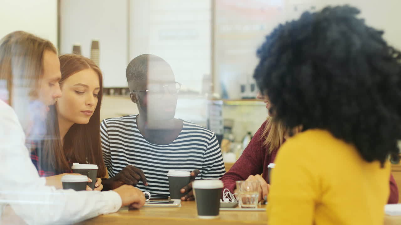 la cámara se enfoca en un grupo multiétnico de amigos a través de la ventana hablando mientras ve algo en una tableta sentada en una mesa en un café