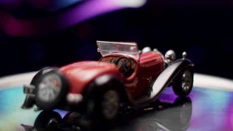 a close-up of a red vintage toy car on a reflective surface with a colorful bokeh background