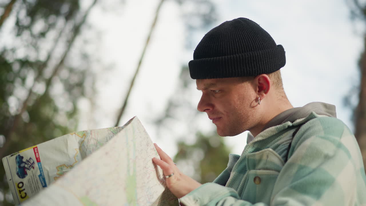 caucasian backpacker studying map near trees, profile view with focused gaze, hands smoothing paper folds, jacket and beanie visible, soft sky through canopy, reflective planning before next leg