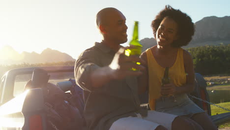 couple with backpacks sitting on tailgate of pick up truck on road trip by lake drinking beer