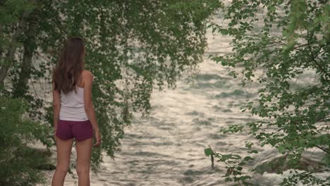 a beautiful european woman looking back over her shoulder and waving at the camera from the edge of a river