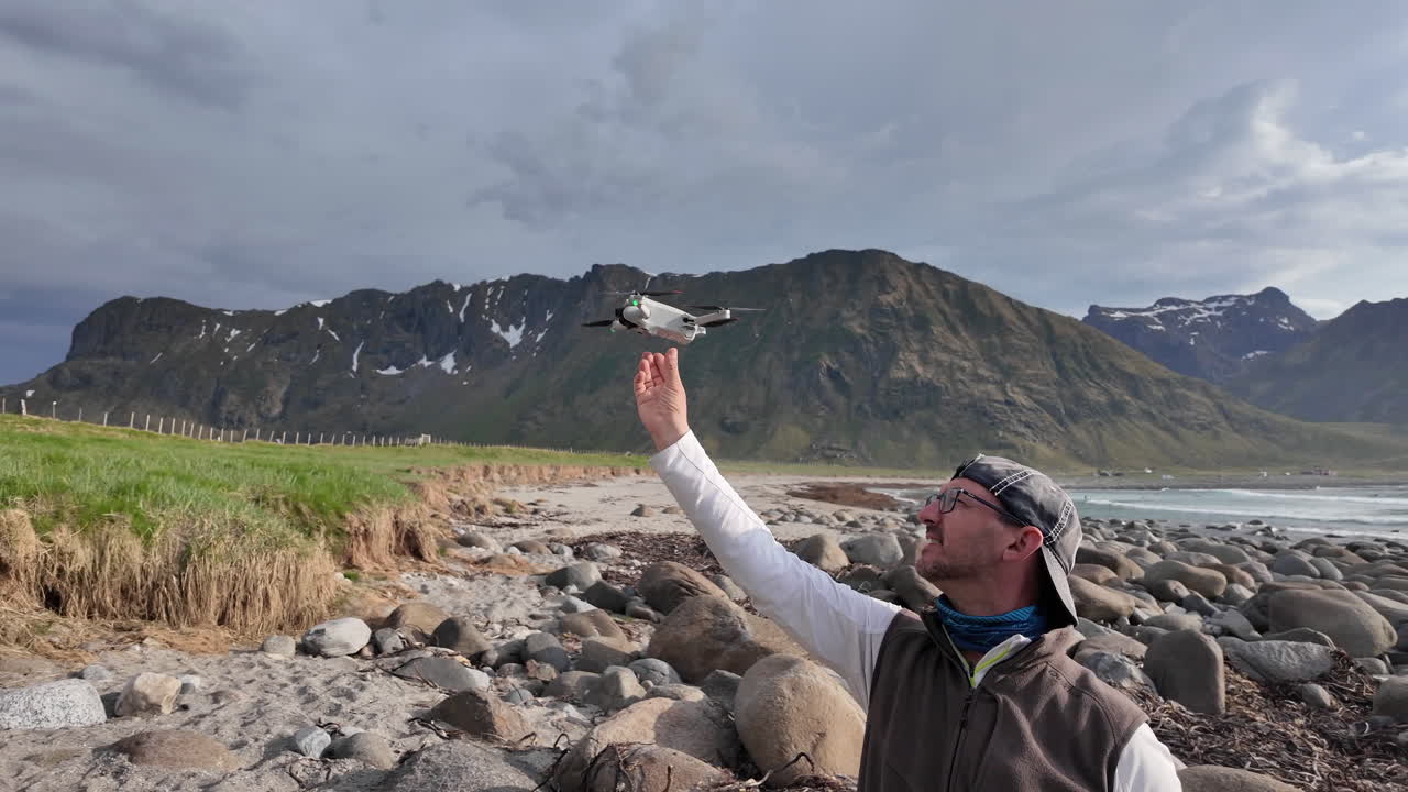 Professional videographer carefully landing remote controlled drone on hand while standing on rocky seashore amid scenic Lofoten Islands landscape with rugged coastline and cloudy sky