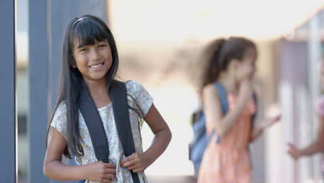 smiling girl with backpack standing outside school, other students in background, copy space