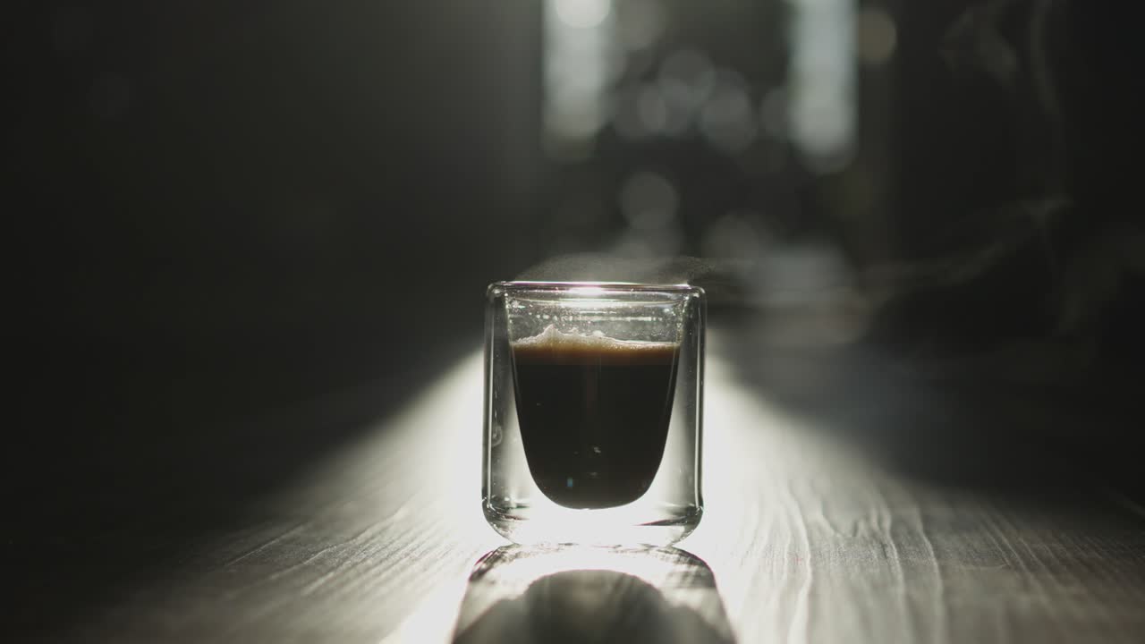 Primo piano di un singolo caffè espresso servito in una tazza di vetro su un tavolo di legno, illuminato da luce naturale con profondità di campo ridotta, creando un'atmosfera suggestiva e cinematografica