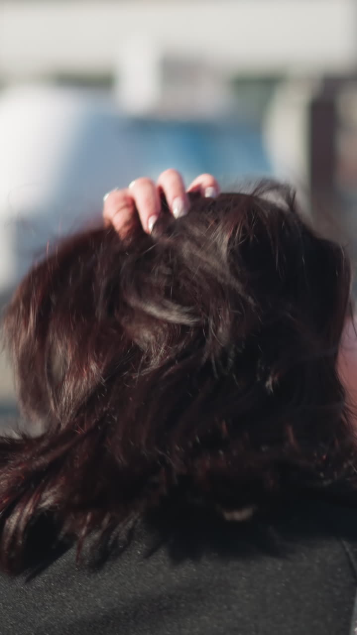 검은 코트를 입은 갈색 머리카락의 여자가 그녀의 머리카락을 조정하고, 왼손에 색 은 손을 과시하며, 빛 도시 거리에서 배경에 건물 흐릿함이 있습니다.