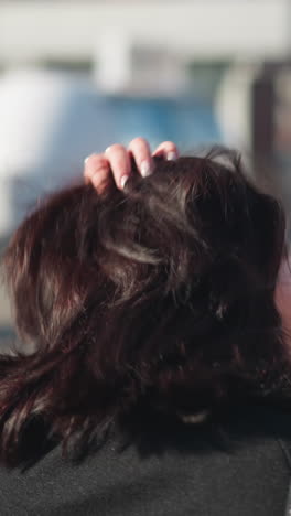 mujer de cabello marrón en un abrigo negro ajusta su cabello, alardeando de uñas blancas pulidas en su mano izquierda, en una calle urbana soleada con el edificio borroso en el fondo