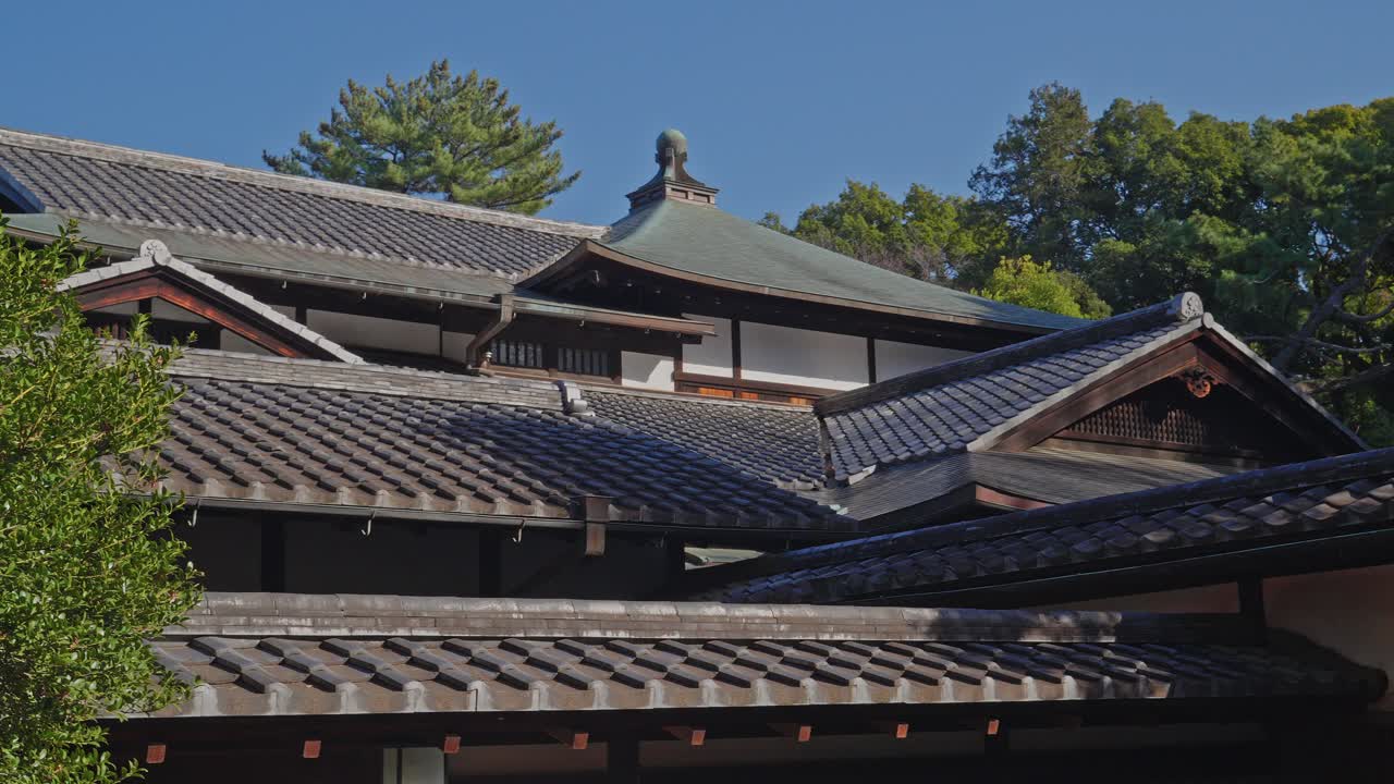 En jævn panorering mod højre over lagdelte lertegltage og de elegante gavle på en traditionel japansk ejendom mod en klar himmel.