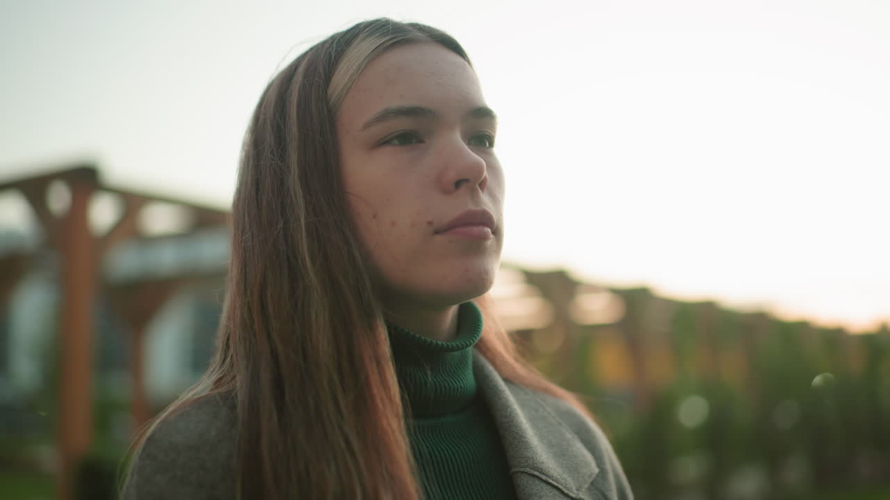 Lady in gray coat gazes thoughtfully into distance outdoors, hair strands swaying gently in breeze, blurred background of residential buildings and wooden structure