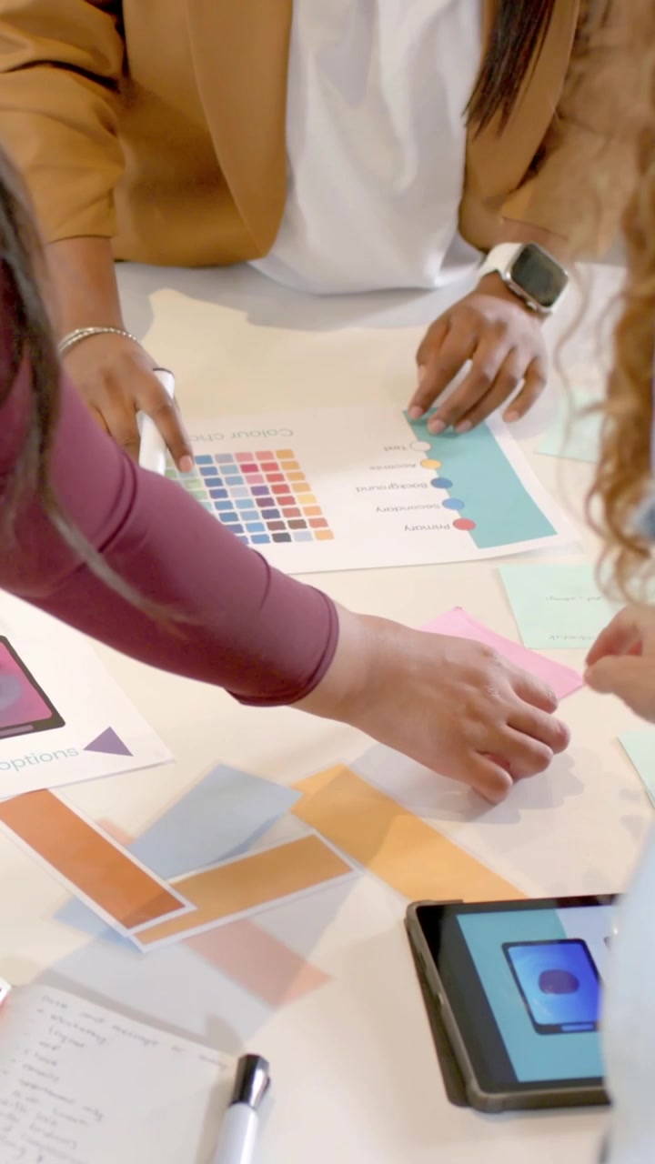 Video verticale: Donna con blazer color cammello che indica e guida il team a un tavolo bianco, posizionando campioni vicino a un tablet