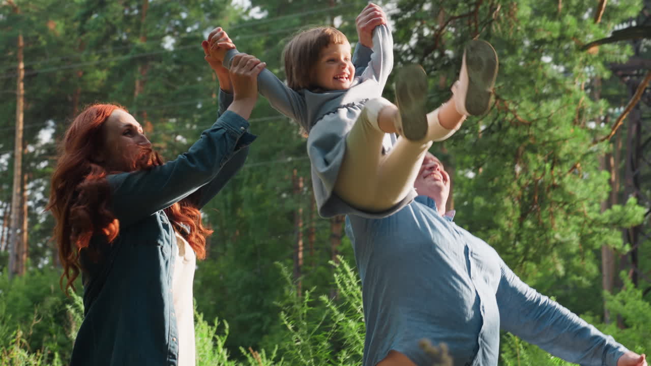 Side view of cheerful parents walking through forest holding hands with smiling daughter as they lift her playfully in sunlight, surrounded by lush greenery, expressing family togetherness outdoors