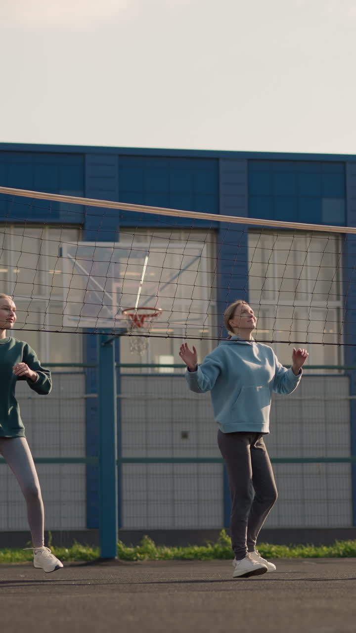 후디를 입은 여성들이 코트 밖에서 배구 훈련을 하고 있으며, 배경에는 농구 허프, 초록색, 건물 등이 있으며, 여성들은 연습 세션 중에 손을 들어뛰고 뛰어다닌다.