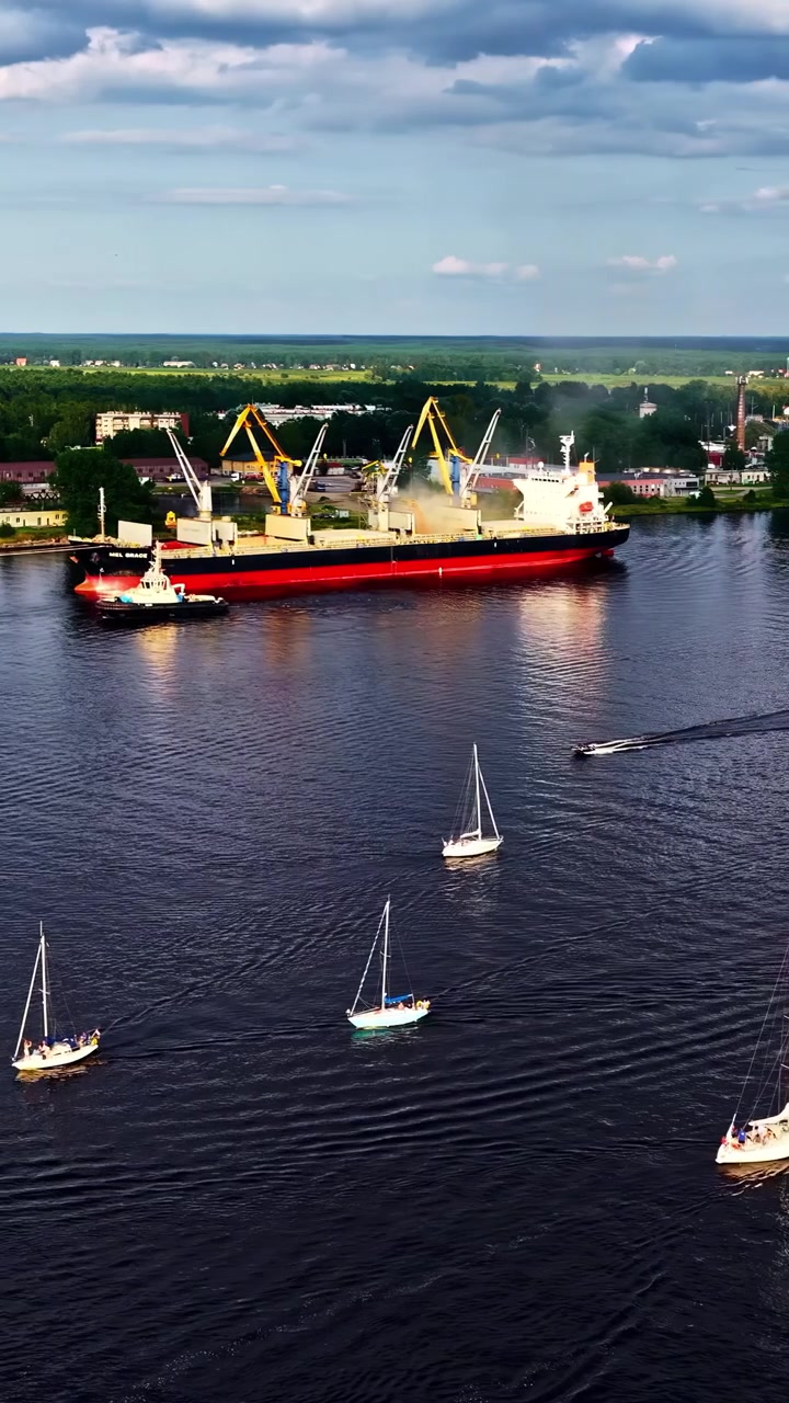 Lastfartyg och segelbåtar rör sig över hamnvattnet under molnig sommarhimmel, vertikal flygbild av sjötransport