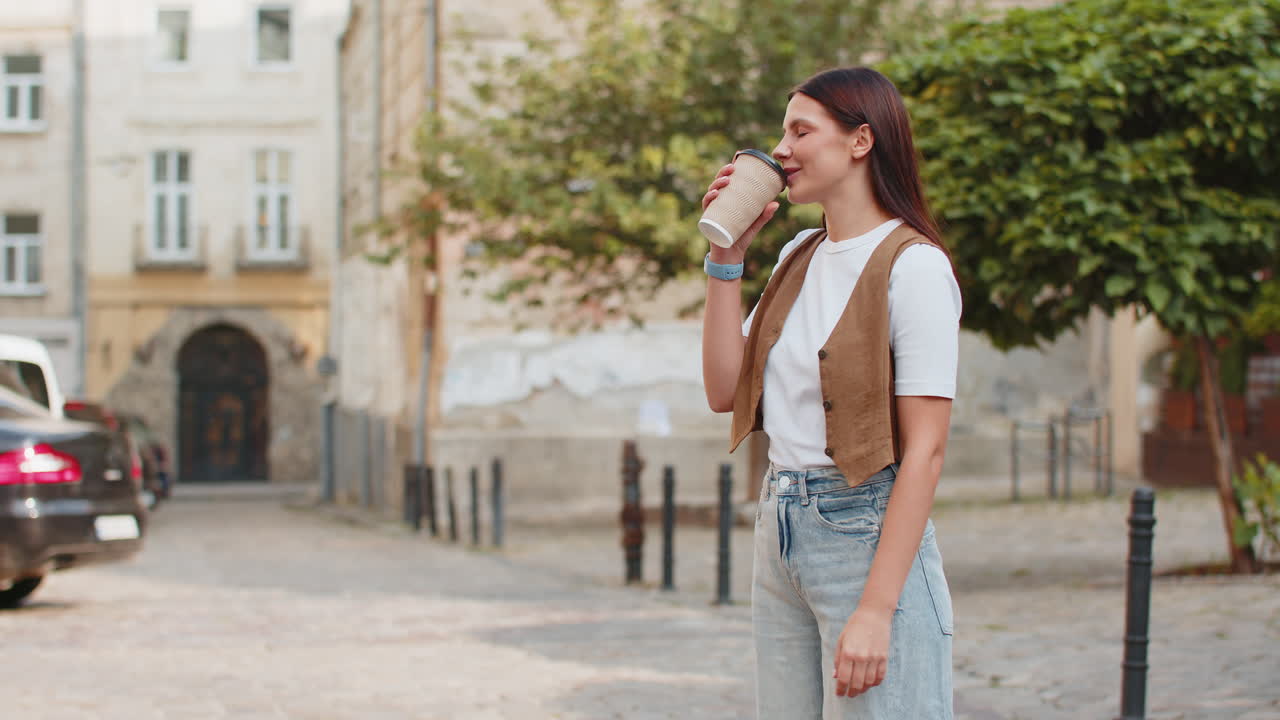 Happy caucasian young pretty woman enjoying morning coffee hot drink and walking on city street