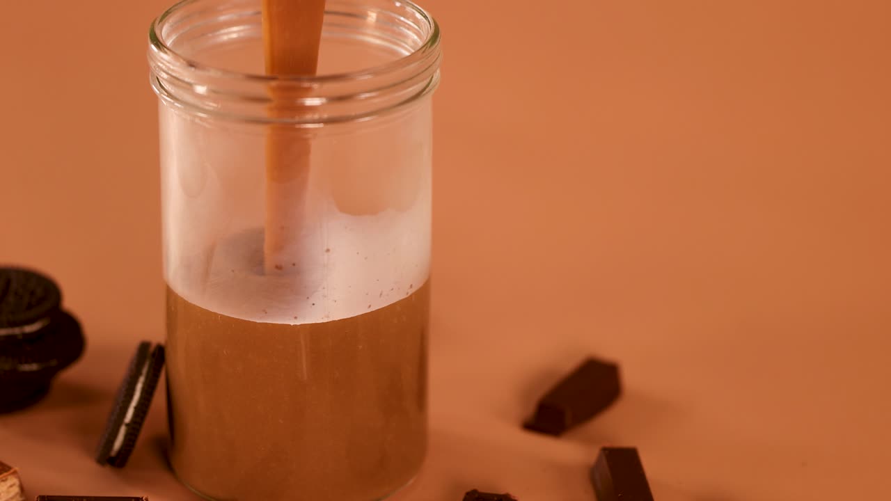 Frullato al cioccolato versato in un barattolo di vetro, circondato da biscotti e cioccolato, con illuminazione calda da studio