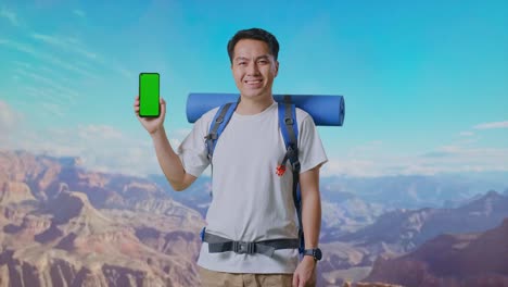 asian male hiker with mountaineering backpack smiling and showing mock up green screen smartphone while traveling at the top of mountain
