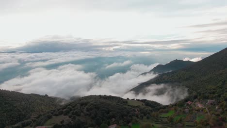 Flight above beautiful village buildings on mountaintop plateau towards puffy white clouds in valley and sun, aerial approach