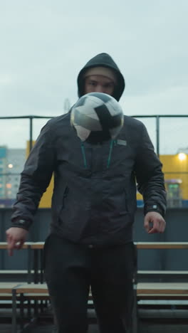 atleta haciendo malabarismos con la pelota con la rodilla durante el entrenamiento en la arena deportiva urbana, demostrando enfoque, contra un paisaje urbano moderno con bancos, edificios y equipos de fitness al aire libre en el fondo