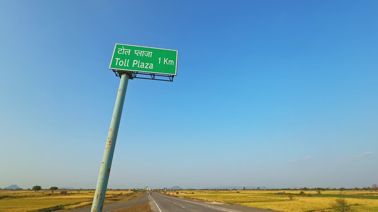 Inquadratura ampia statica di un cartello autostradale verde "Casello 1 Km" montato su un palo alto, che si affaccia su una lunga strada dritta che attraversa campi agricoli dorati sotto un cielo azzurro e limpido.