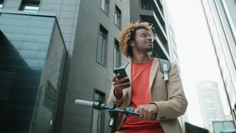 african american man standing with e-scooter in city and using smartphone