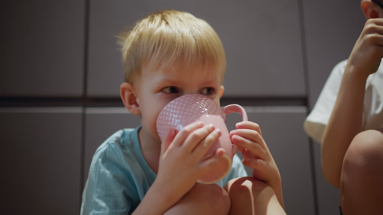 Young boy in light blue shirt sitting on floor holding pink cup with both hands, drinking with focused expression in bright kitchen setting, portraying everyday childhood moment of refreshment