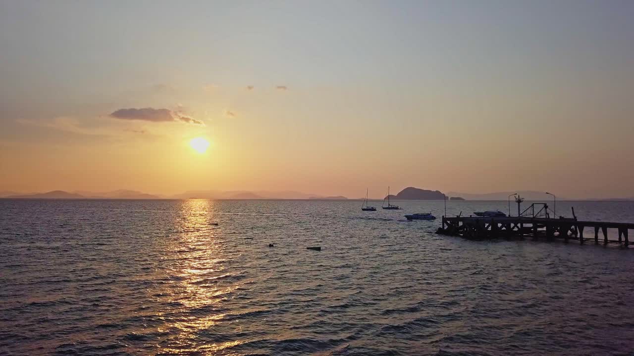A serene sunset view at Koh Yao, Thailand. The golden sun sets over calm ocean waters, casting warm reflections, with distant islands, boats, and a pier creating a tranquil seascape.