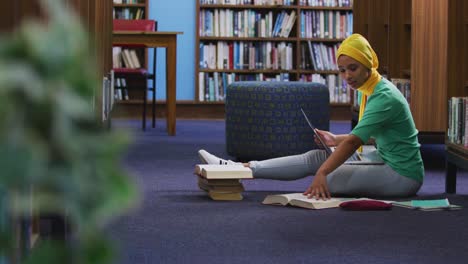 estudiante asiática vestida con un hijab amarillo sentada con un libro abierto y usando una computadora portátil