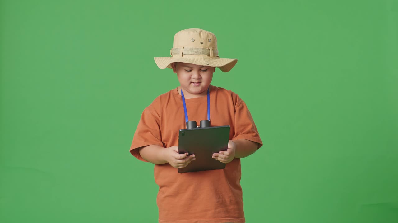 모자와 안경을 입은 아시아 관광 소년은 태블릿을 사용하고 녹색 화면 배경에 서서 미소 짓고 있습니다. 소년 연구자는 여행, 관광, 모험 개념의 무언가를 조사합니다.
