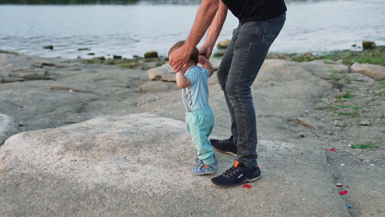 Man wearing jeans leading his little son studying to walk. Beautiful baby boy in blue clothes stepping by stones. Bank of the river setting.