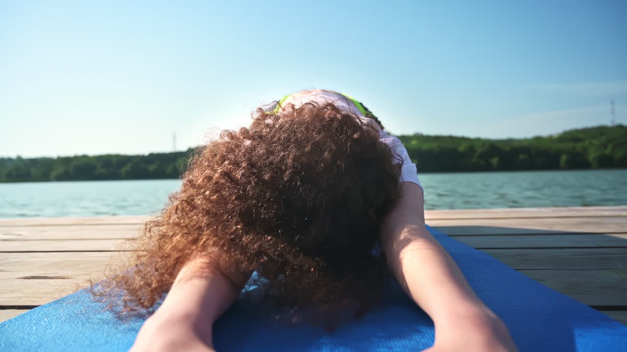 Young curly woman practicing yoga near the lake while wearing black protective mask. Sunny day. Meditating in child's pose on the yoga mat. Corona Virus idea. Chisinau, Moldova