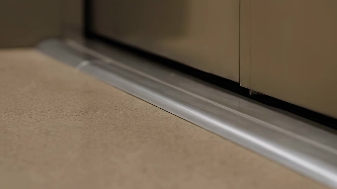 Una vista da vicino al suolo mostra una soglia in alluminio accanto a un pavimento texturizzato, con un morbido riflesso in movimento lungo il bordo metallico arrotondato, dettagli interni puliti e toni neutri e smorzati in un tranquillo ambiente residenziale.