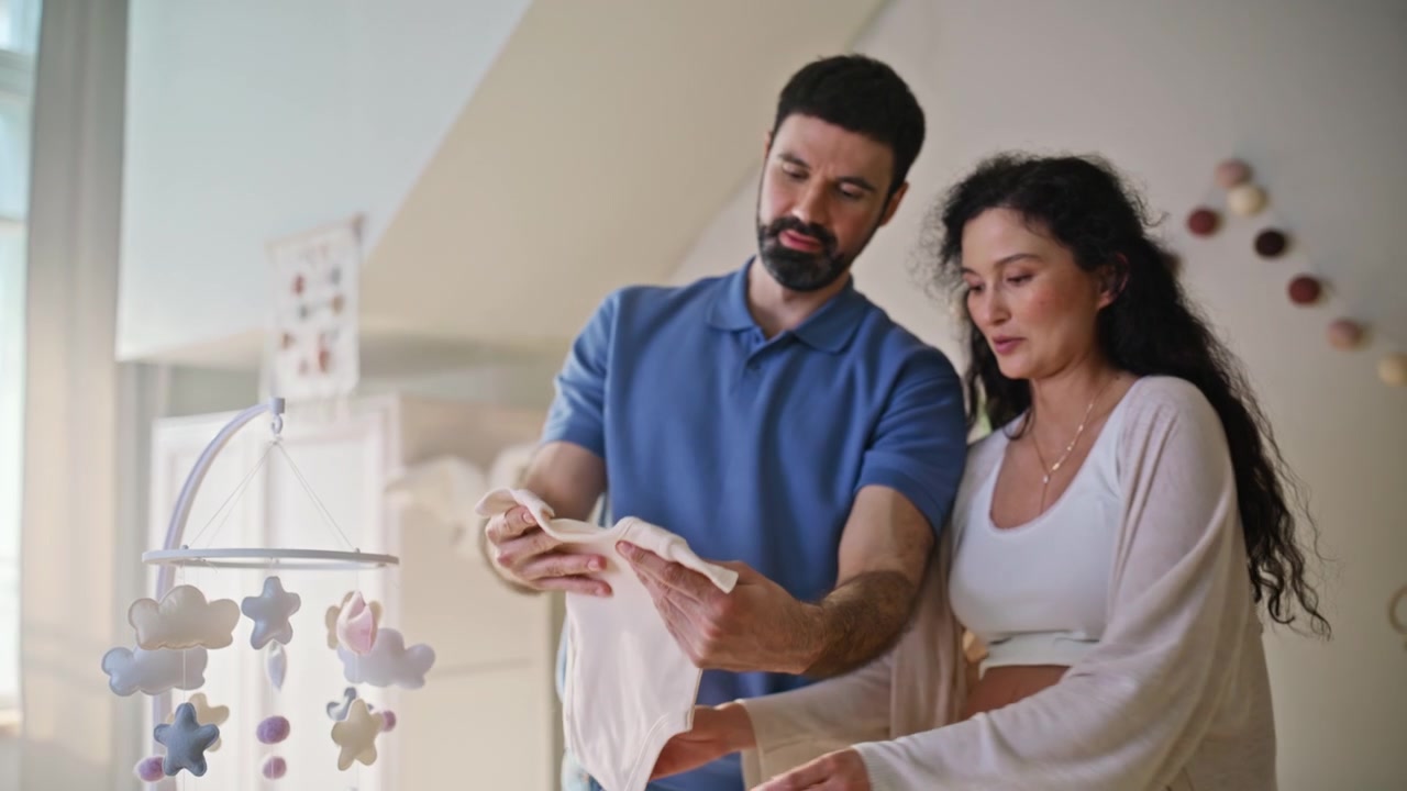 Expecting couple arranging baby clothes nursery. Man woman preparing bodysuit.