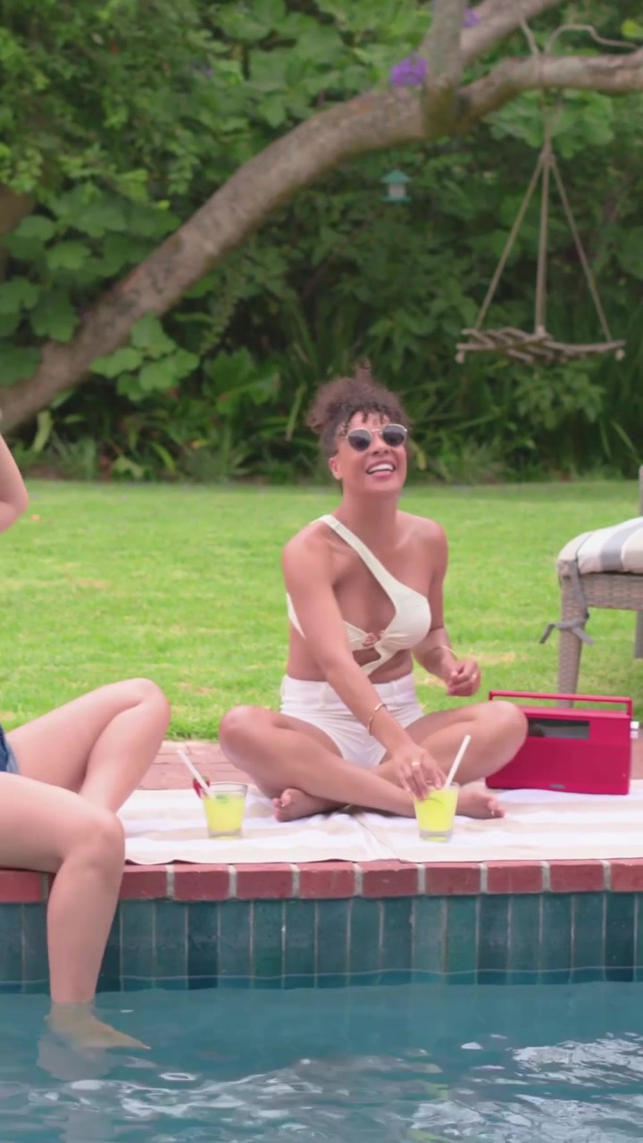Vertikales Video: Tanzende Frau in weißem Anzug nippt am Pool aus einer gelben Tasse, zu Musik aus einem roten Lautsprecher.
