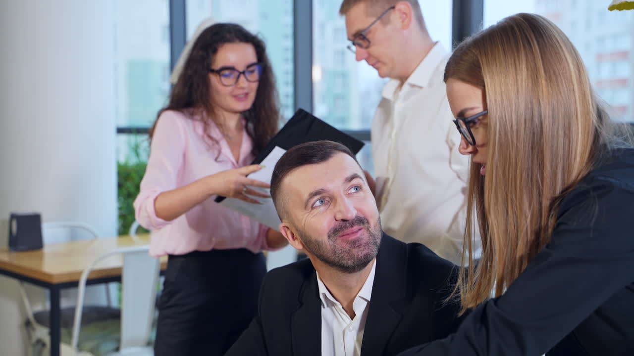 Businessman sitting at computer and listening to his female employee talking. Couple employees standing and talking about the charts at the backdrop.