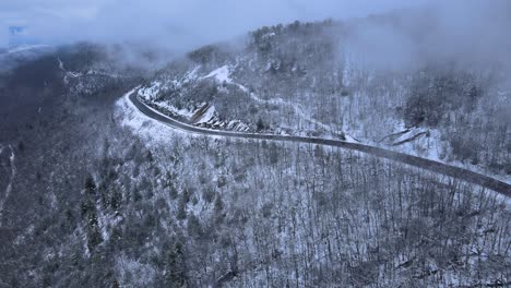 Flying-through-clouds-over-snow-covered-mountains-and-forests-in-a-mountain-valley-with-a-scenic-highway-below,-on-a-cloud-winter’s-day