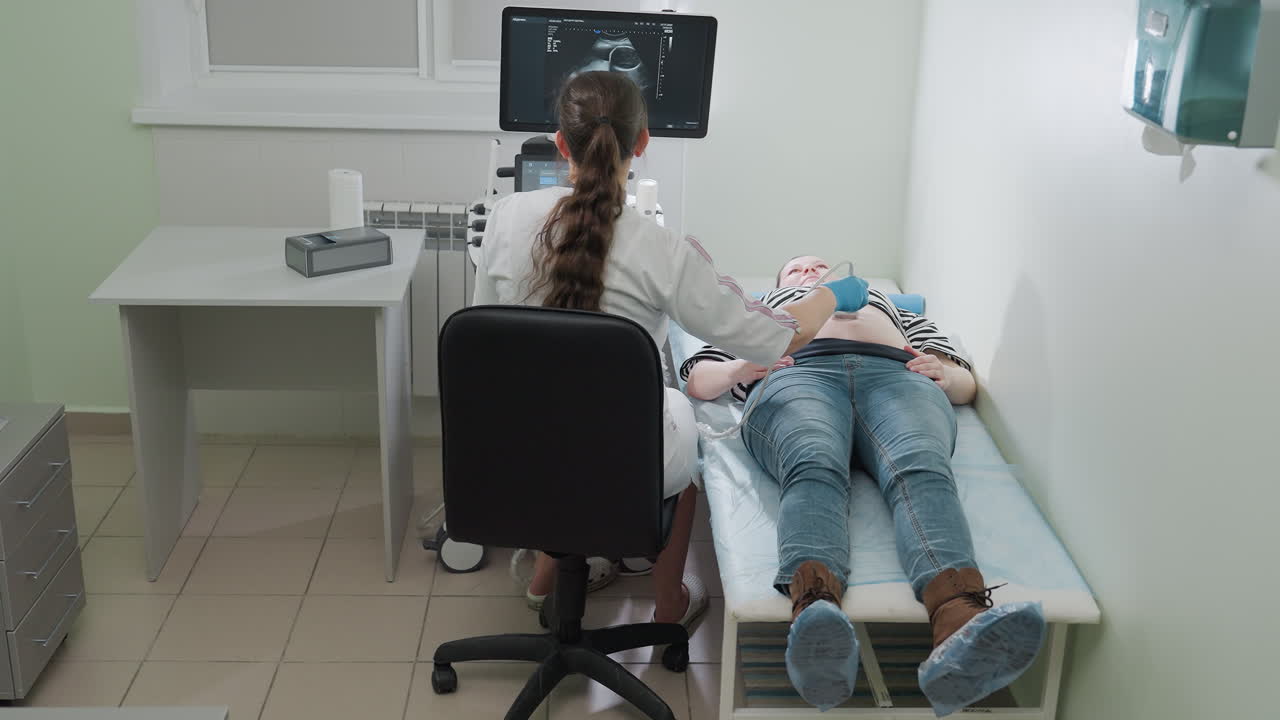 Female gynecologist in clinical setting examines abdomen of female patient lying on hospital bed while focusing on monitor displaying ultrasound image with wardrobe and table visible in workspace