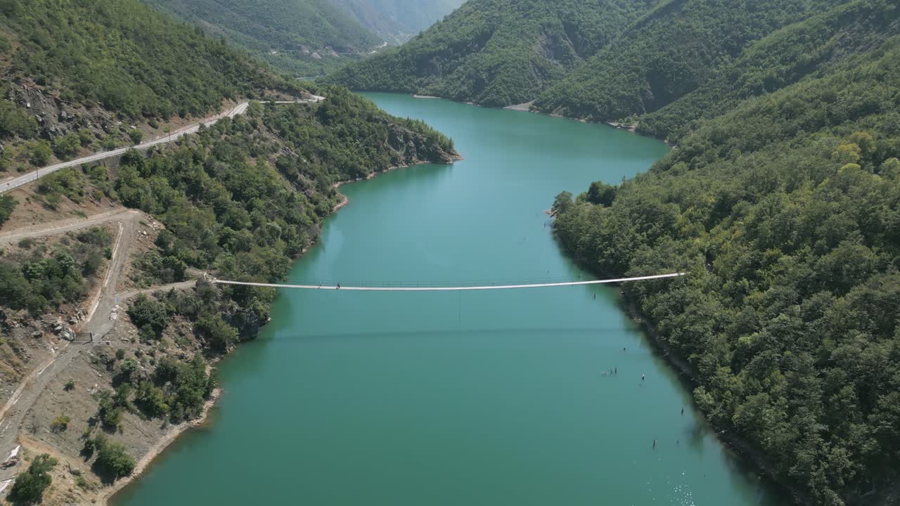 Pedestrians cross a long suspension bridge over turquoise waters in a lush mountain valley