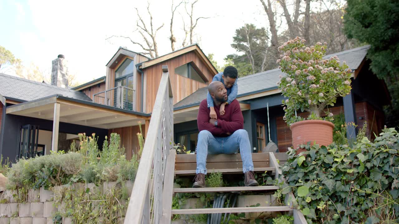 African American father sitting on wooden stairs at home as son climbing behind into piggyback hold
