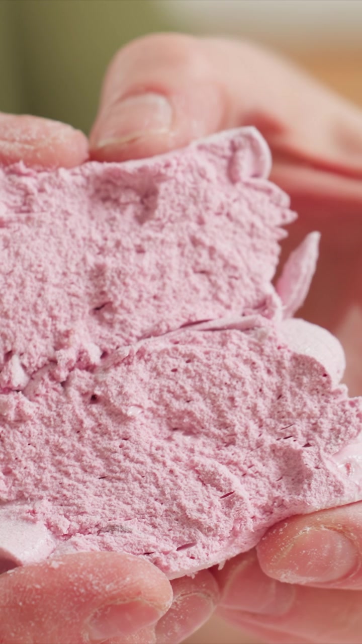 Primo piano di mani che tengono un materiale rosa poroso, rivelandone la friabilità. Le mani manipolano un oggetto morbido con crepe visibili sulle dita. Lo sfondo sfocato enfatizza la messa a fuoco sul soggetto.