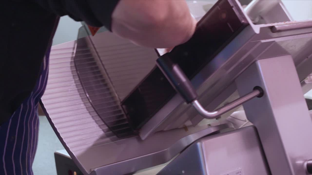 제복을 입은 델리 작업자는 산업용 슬라이서를 사용하여 델리 고기와 치즈를 준비합니다.