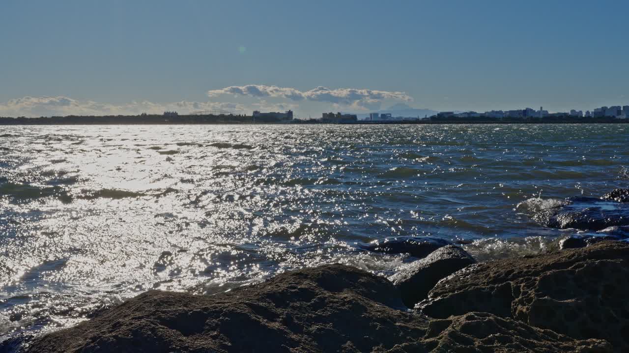 A bright, shimmering reflection of sunlight on the dark, choppy bay water, with rugged rocks framing the bottom foreground